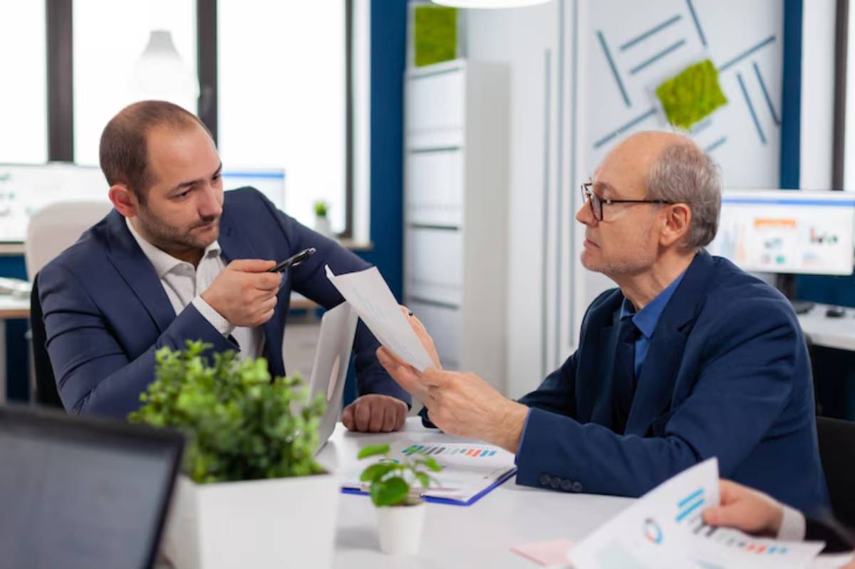 Two men in business attire discuss documents at a modern office table, with laptops and charts visible in a bright, professional environment.