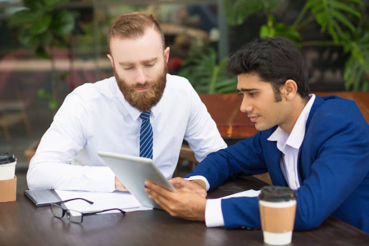 Two professionals discuss over a tablet at a cafe table, with coffee cups and documents visible in a greenery-filled setting.