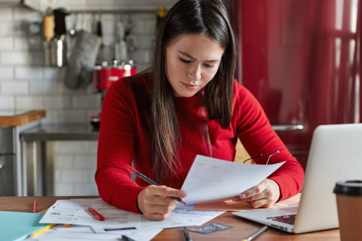 A person in a red sweater reviews documents at a table, surrounded by papers and a laptop in a modern kitchen setting.