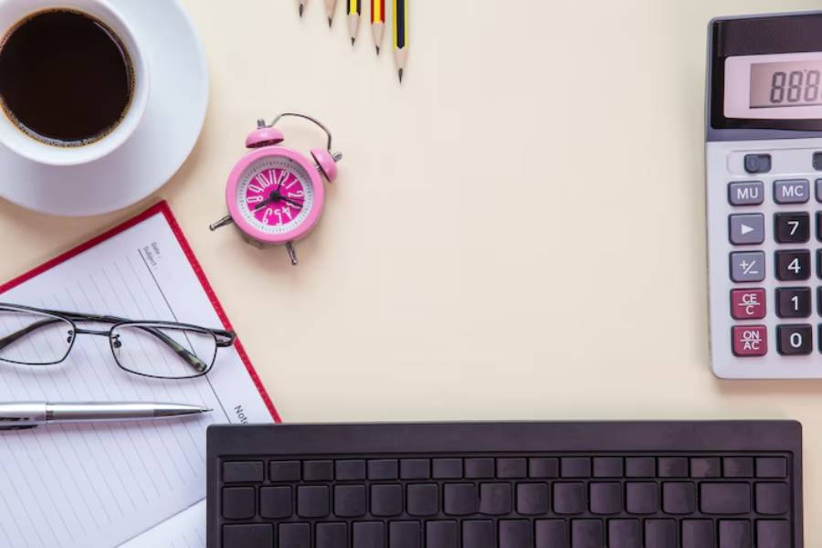 A workspace scene featuring a cup of coffee, pink alarm clock, glasses, notebook, pen, black keyboard, and a calculator.