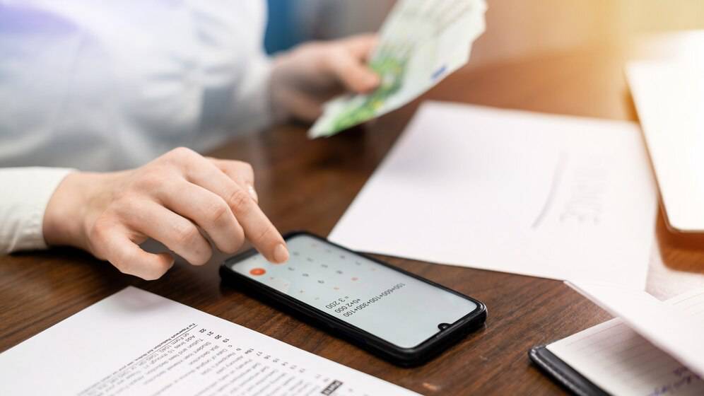 A person uses a smartphone while holding cash, with paperwork laid out on a wooden table.
