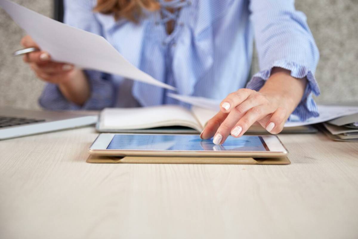 A woman in a blue striped shirt reviews documents while using a tablet on a desk, with a laptop and notepad nearby.