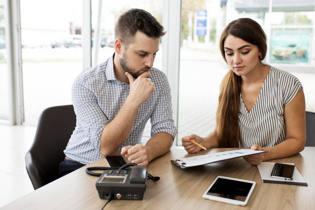 A man and a woman engage in a discussion at a table, reviewing documents and charts with a phone and tablet nearby.