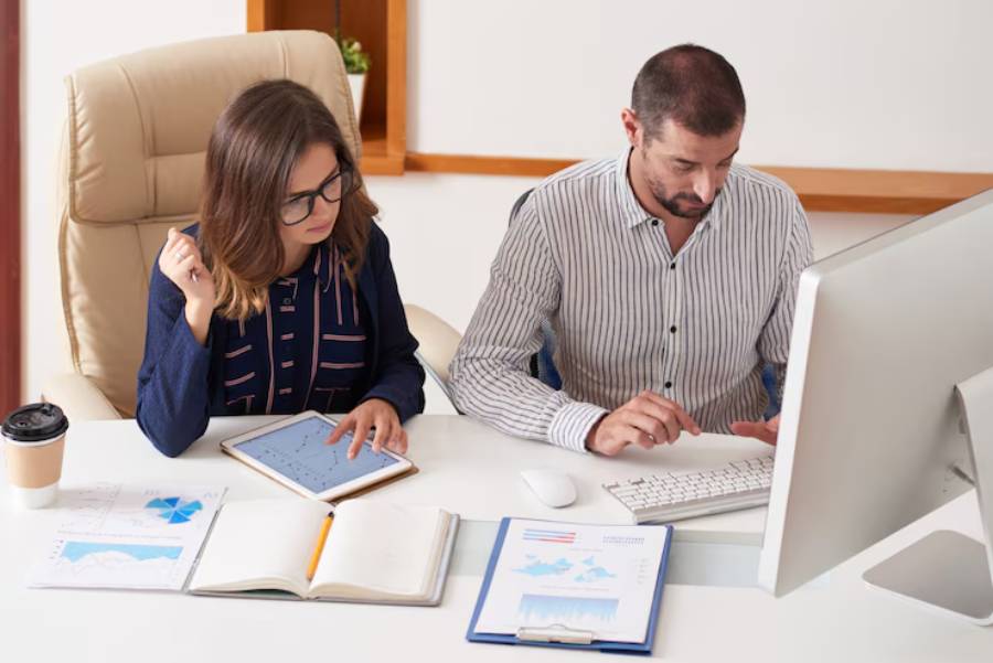 Two professionals engage in a discussion at a desk, analyzing charts on a tablet and computer, surrounded by documents and coffee.