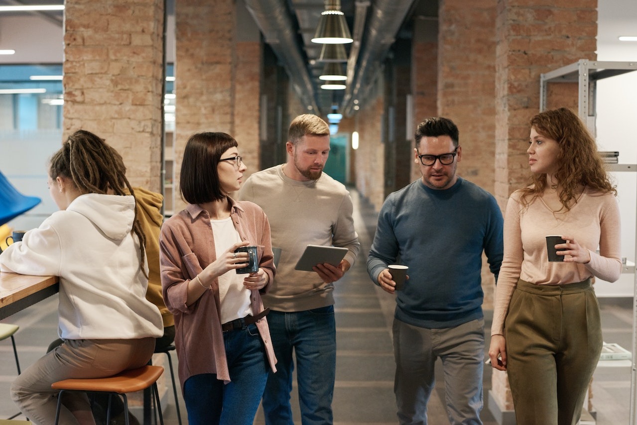 A diverse group of five people chatting and walking in a modern office space with brick walls and industrial design features.