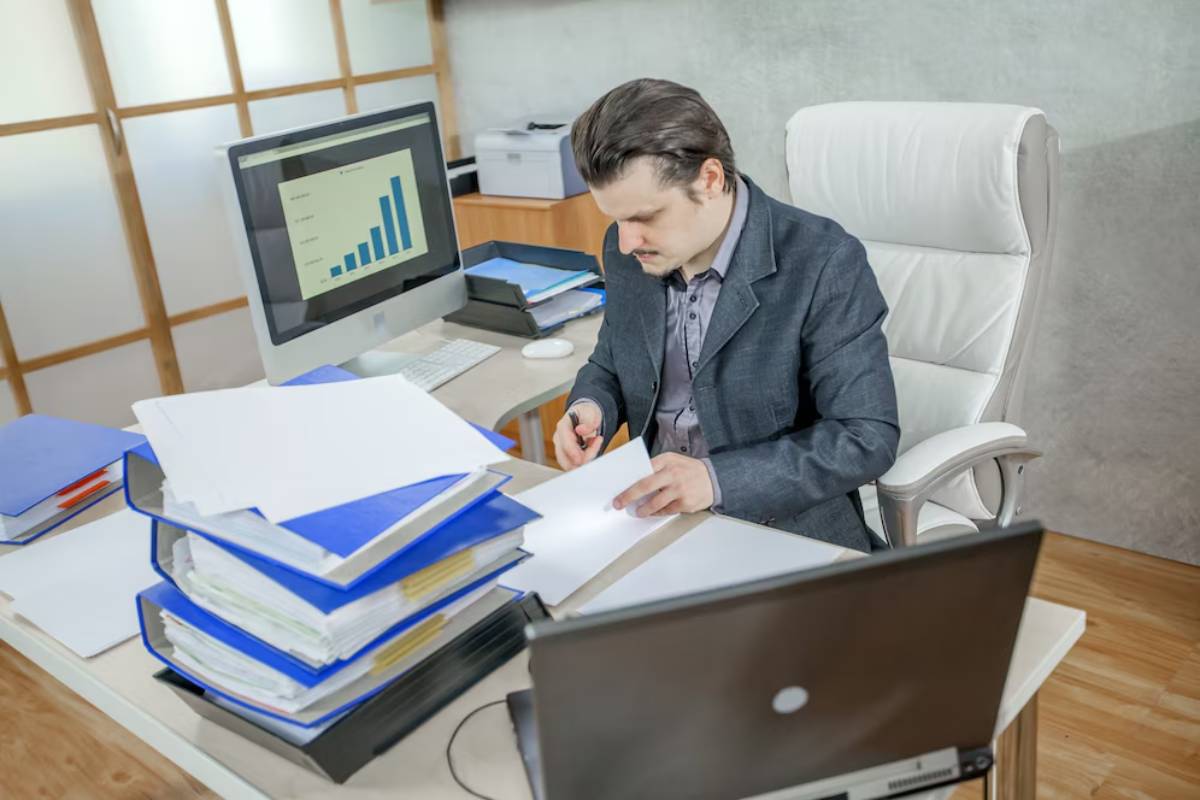 A professional in a suit sits at a desk with a computer, analyzing data on a screen, surrounded by paperwork and organized files.