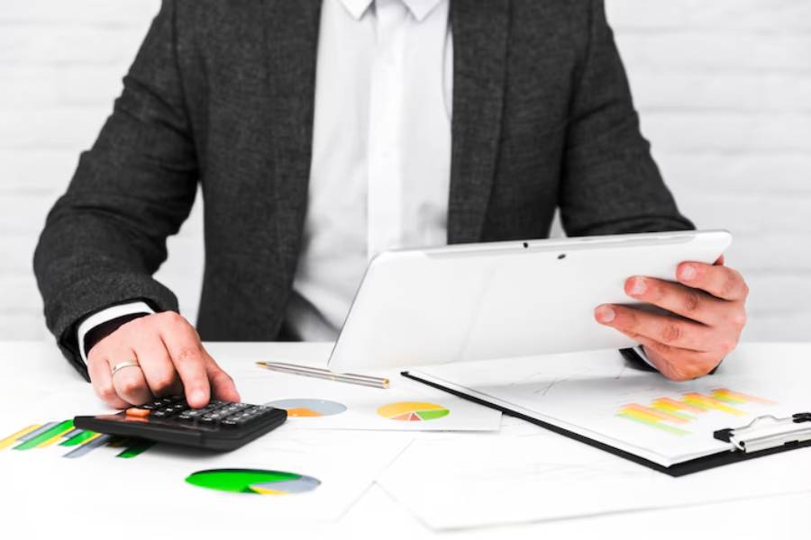 A businessman in a suit using a calculator and a tablet while analyzing colorful charts on a desk.