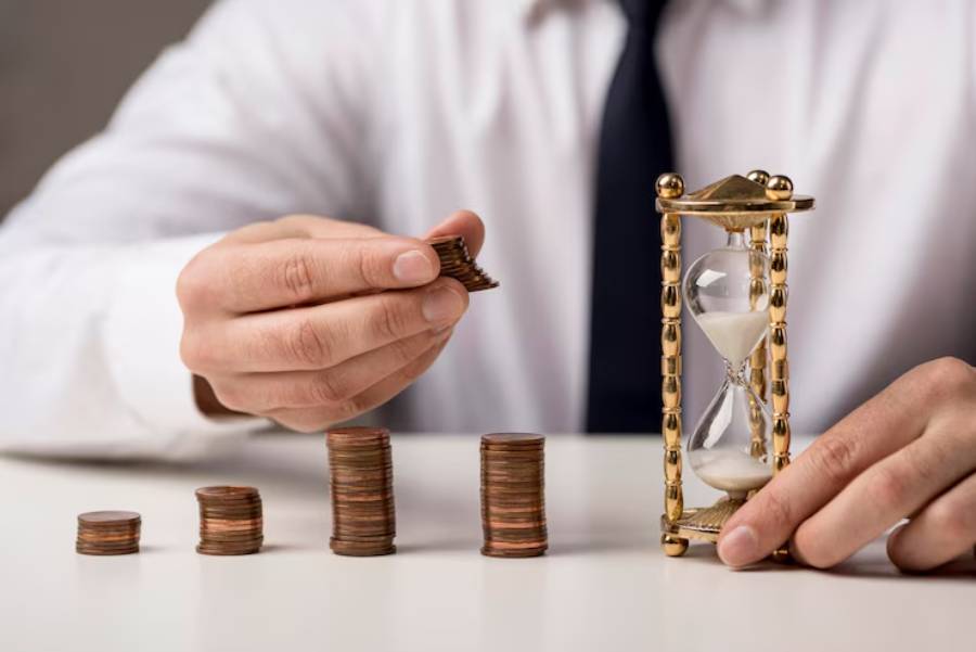 A person in a white shirt and tie stacks coins beside an hourglass, symbolizing the balance of time and money.