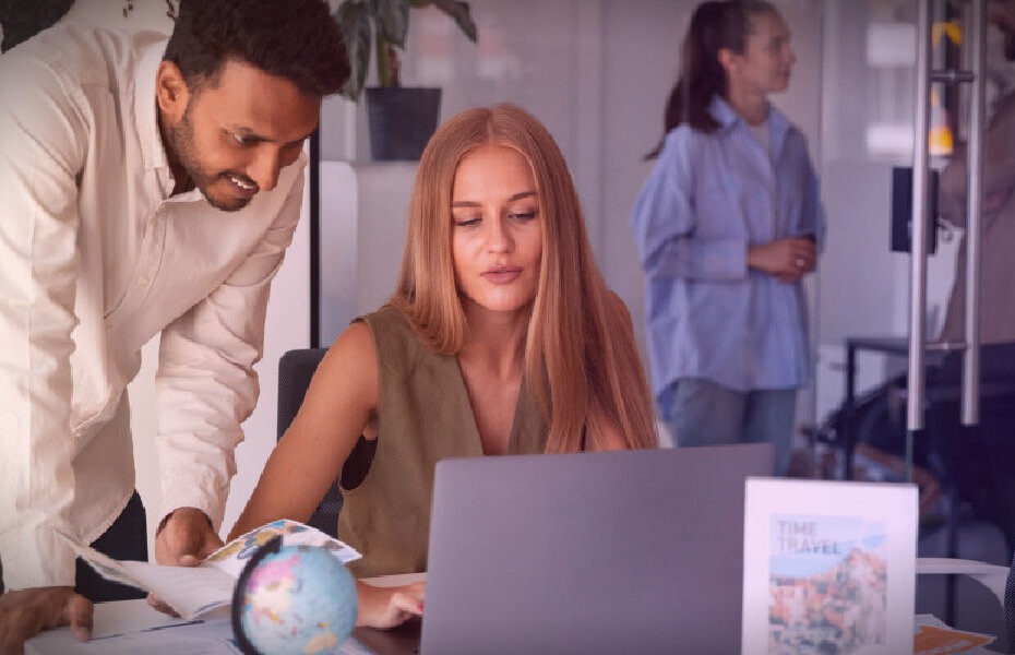 A woman with long hair sits at a desk, engaged with a laptop and documents, while a man leans in to discuss. A globe is nearby.