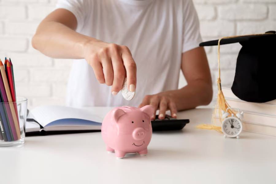 A person deposits a coin into a pink piggy bank on a desk, with a graduation cap, clock, notebook, and colored pencils nearby.