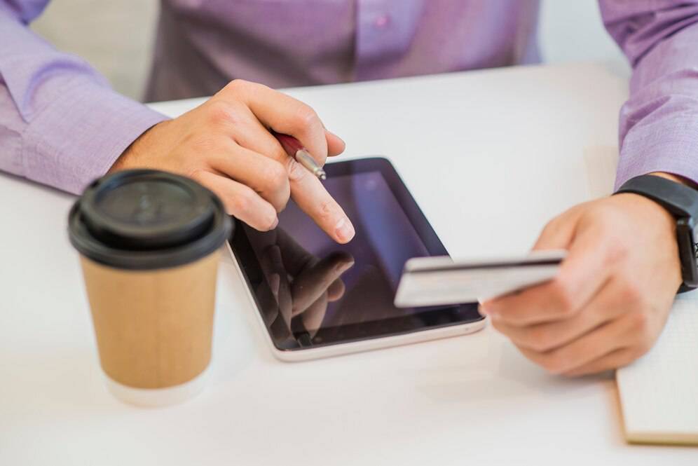 A close-up of a person's hands using a tablet and holding a credit card, with a coffee cup nearby on a table.