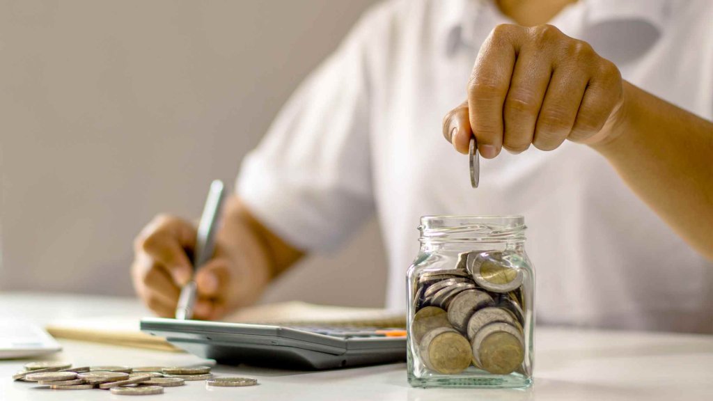 A person drops a coin into a glass jar filled with coins while writing notes and using a calculator on a desk.
