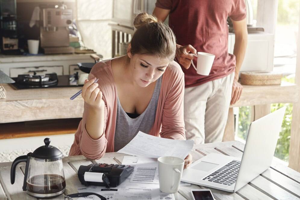 A person analyzes paperwork at a wooden table, accompanied by a laptop, coffee pot, and another individual holding a cup.