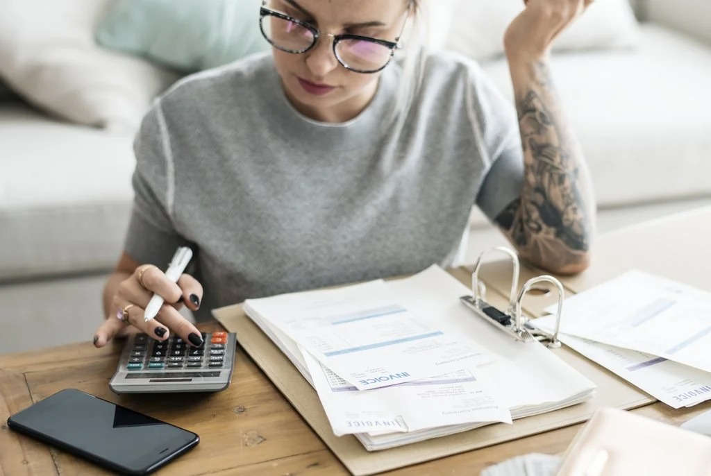 A person with a tattoo calculates expenses using a calculator, surrounded by invoices and a smartphone on a wooden table.