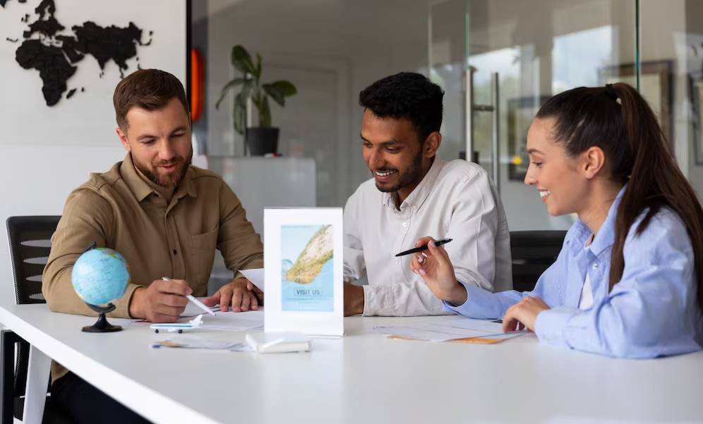 Three individuals engaged in a discussion at a modern office table, with a globe and promotional material visible.