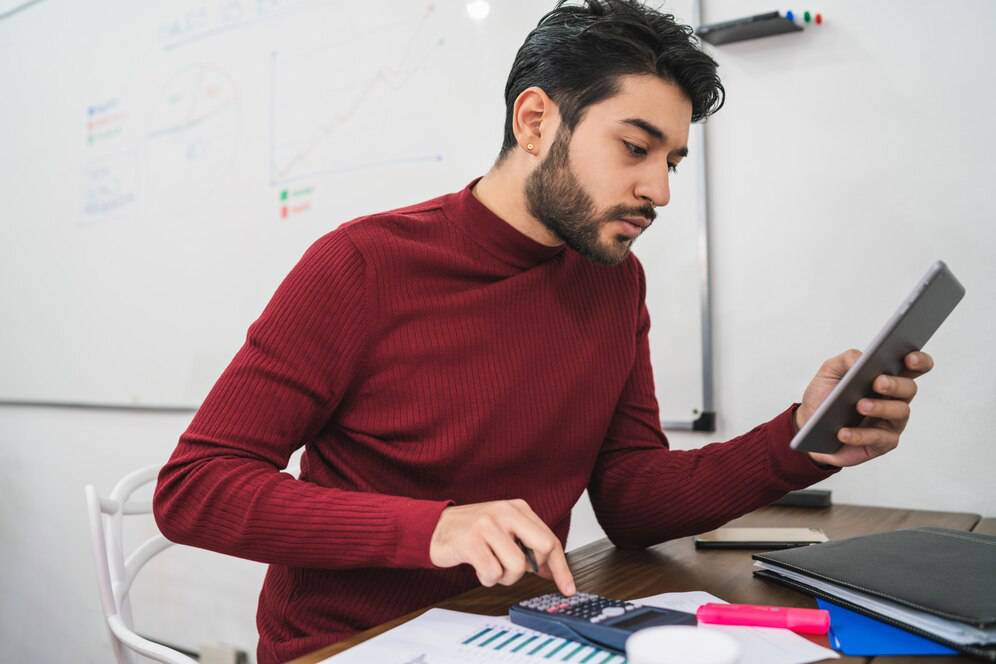 A person in a red sweater uses a calculator and tablet at a desk with documents, while a whiteboard with graphs is visible behind.