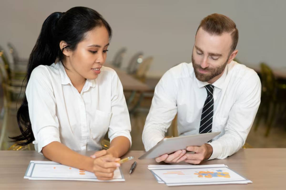 A man in a white shirt and a tie shows a tablet to a woman in a white blouse, surrounded by documents and a blurred background.