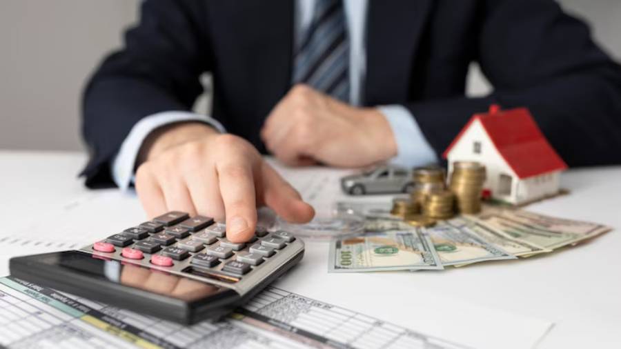 A man calculates finances with a calculator, surrounded by cash, coins, a toy house, and a car model on a document-filled desk.