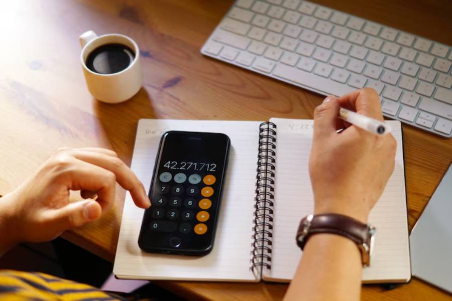 A person calculates numbers on a smartphone calculator beside a spiral notebook and coffee cup on a wooden desk.