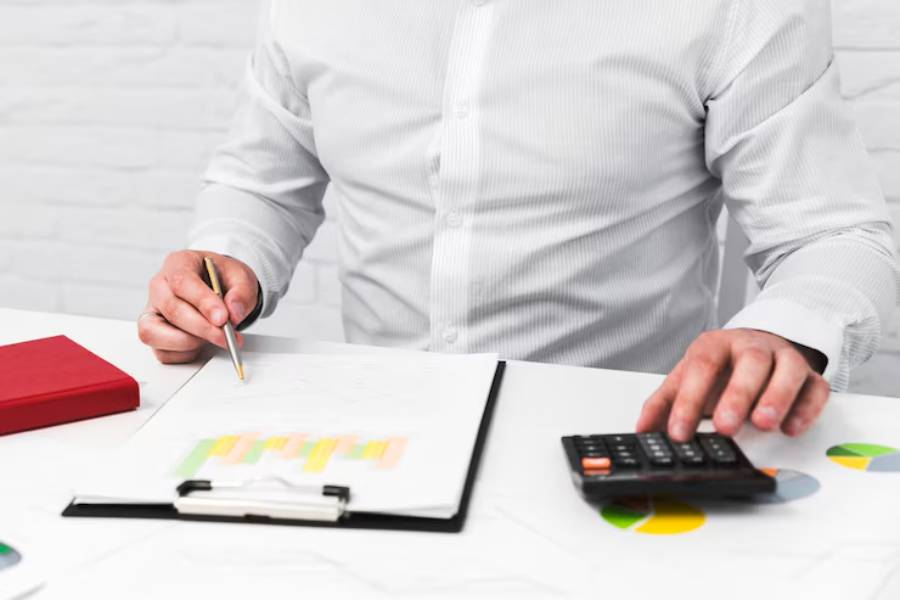 A person in a striped shirt writes on a clipboard while using a calculator, surrounded by charts and a red notebook.