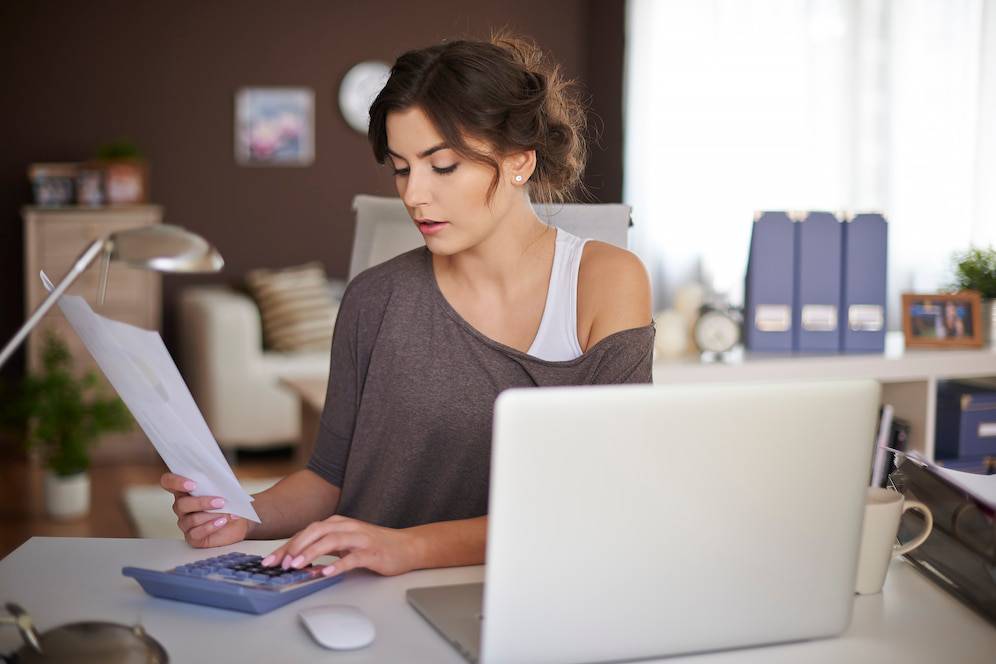 A person in a casual outfit sits at a desk, reviewing documents and using a calculator, surrounded by office supplies and a laptop.