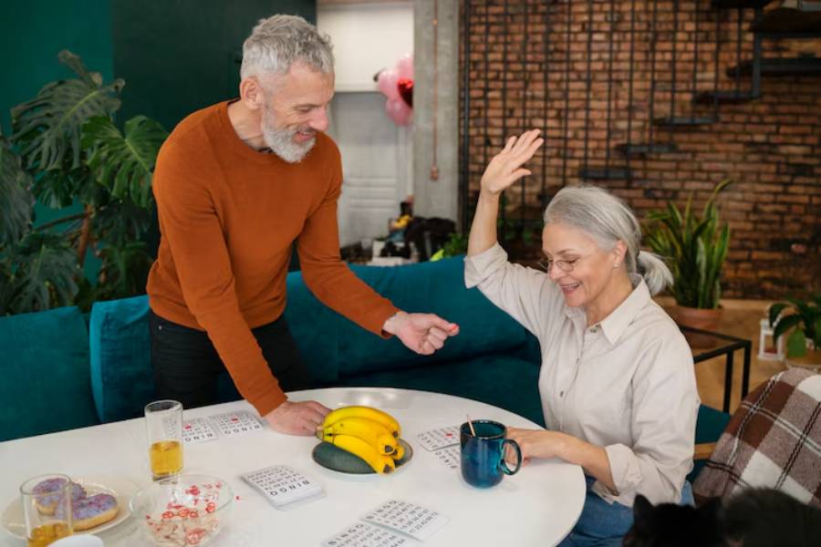 A man passionately gestures while a woman raises her hand in response, surrounded by a cozy setting with plants and games on the table.