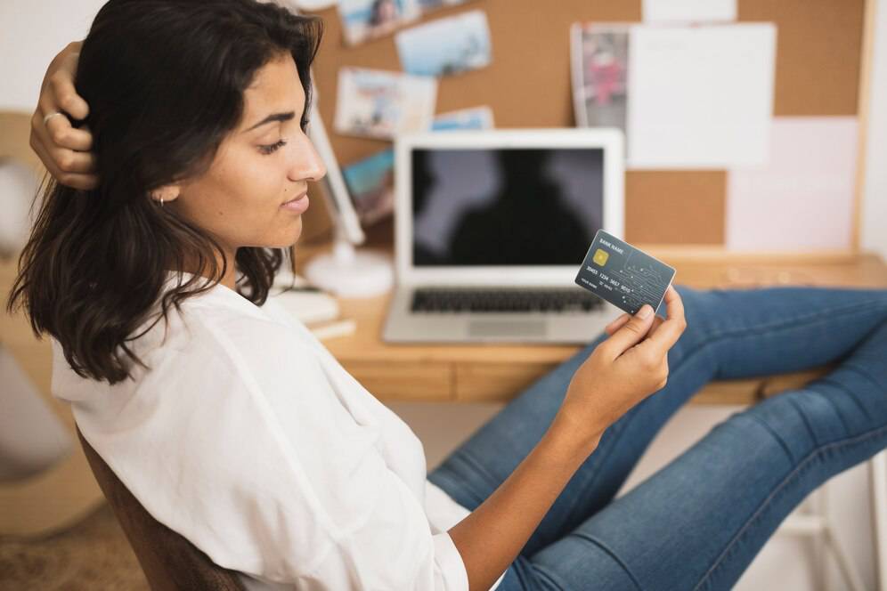 A person sitting at a desk holds a credit card, with a laptop and pinned notes in the background, in a cozy indoor workspace.
