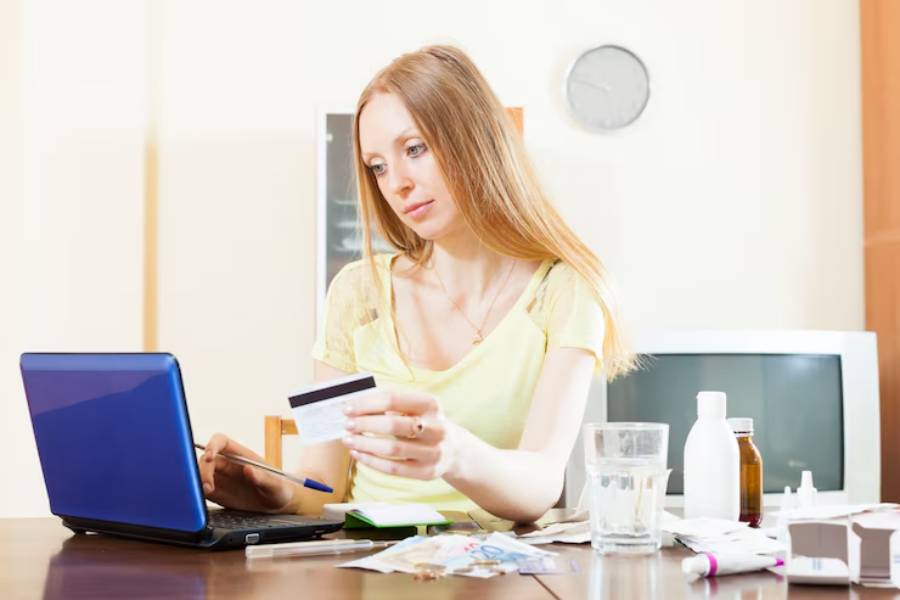 A woman sits at a table with a laptop, holding a credit card and pen, surrounded by medications and a glass of water.