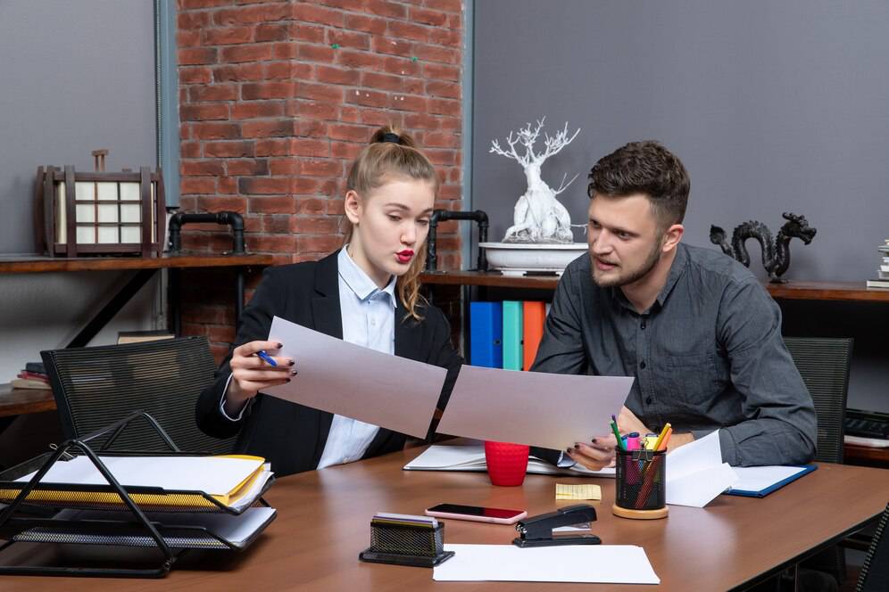 Two professionals are discussing documents at a wooden table, surrounded by office supplies and a brick wall backdrop.