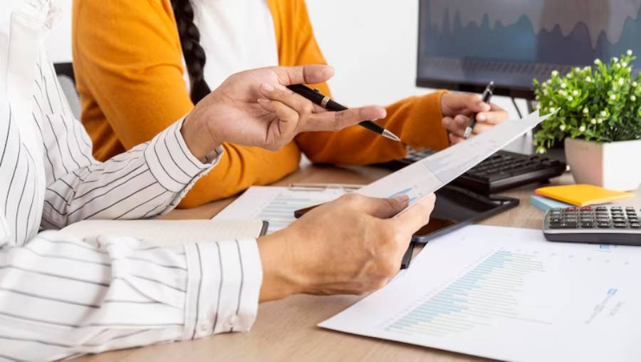 Two professionals collaborate at a desk, discussing printed reports and charts, with a computer and office supplies in the background.