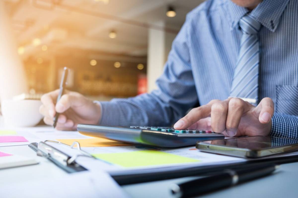 A professional in a plaid shirt calculates figures with a calculator, surrounded by notes, a pen, and a smartphone on a desk.
