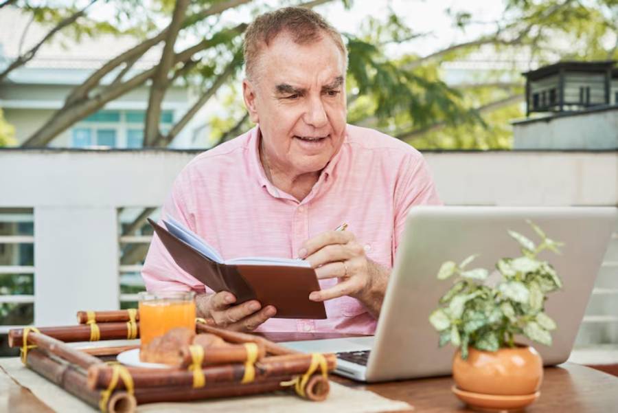 A person in a pink shirt writes in a notebook while seated at a table with a laptop, juice, and a potted plant outdoors.