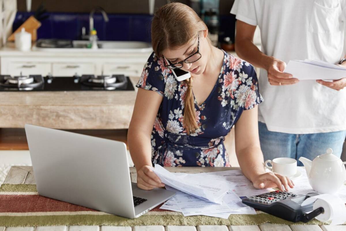 A woman reviews papers at a table with a laptop, calculator, and a teapot while a man stands nearby holding documents.