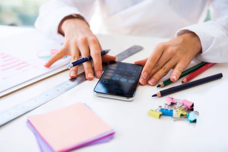 A person using a smartphone with a calculator app, surrounded by stationery items like pens, colored pencils, and sticky notes on a desk.
