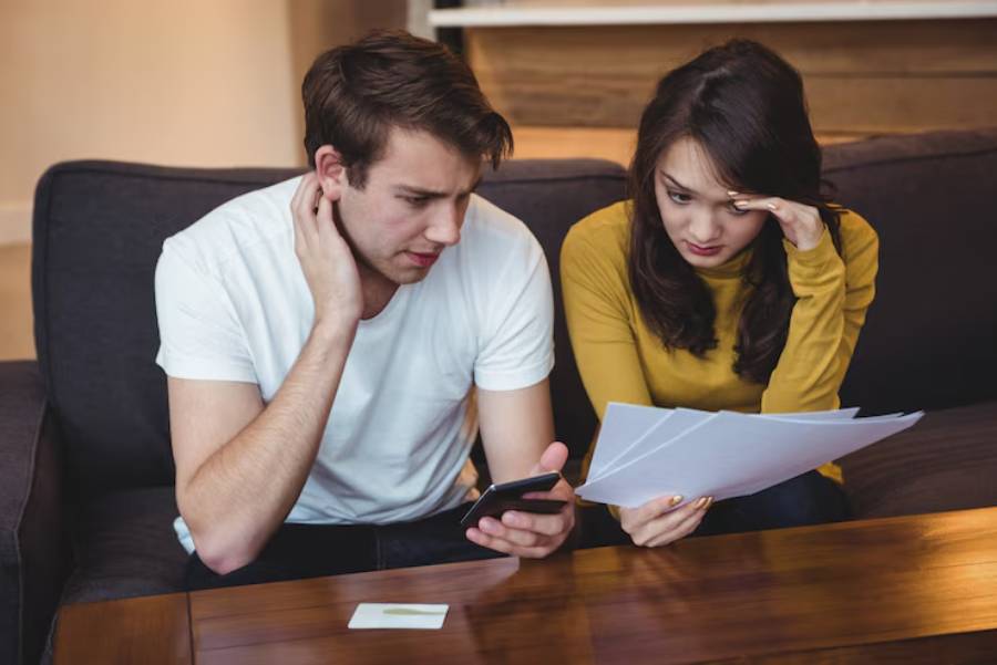 A man in a white t-shirt and a woman in a yellow sweater sit on a couch, reviewing papers while looking at a phone.