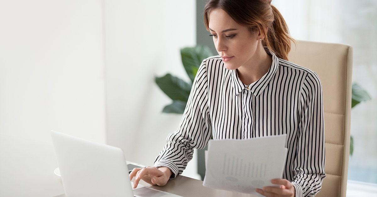 A person in a striped shirt reviews documents while working on a laptop at a bright, minimalistic desk with a plant in the background.
