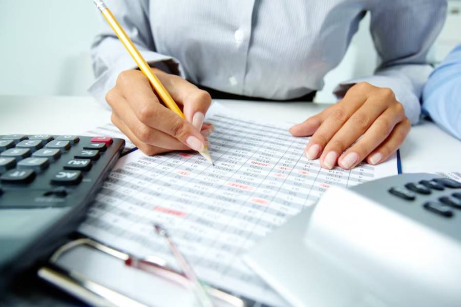 A person in a striped shirt uses a pencil to write on a financial spreadsheet, surrounded by calculators and office supplies.