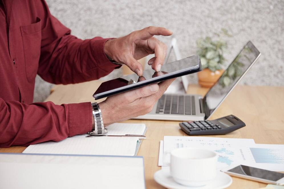 A person in a red shirt uses a tablet while seated at a desk with a laptop, calculator, notebook, and a cup of coffee.