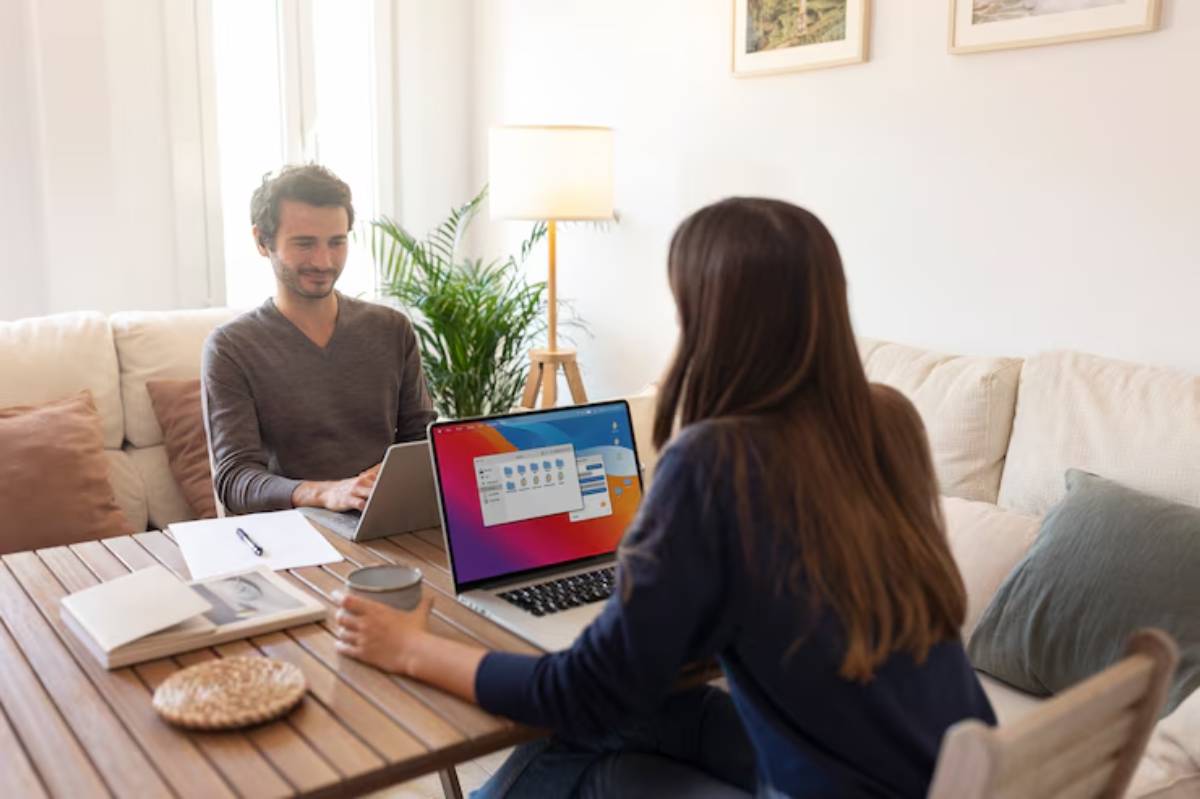 A woman sits at a wooden table with a laptop, holding a cup, engaging with a man on another laptop in a cozy living room.