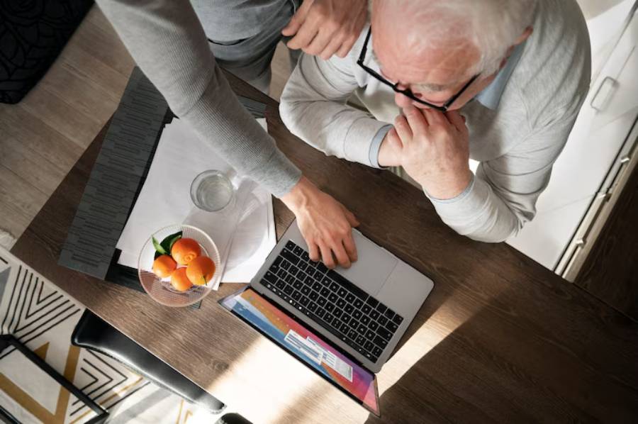 An overhead view of two people interacting at a wooden table with a laptop, a glass of water, and a bowl of oranges.