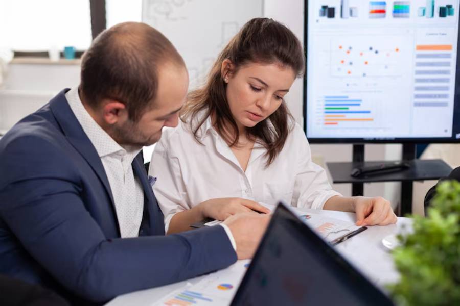 Two professionals collaborate over documents at a meeting, with graphs displayed on a screen in the background.