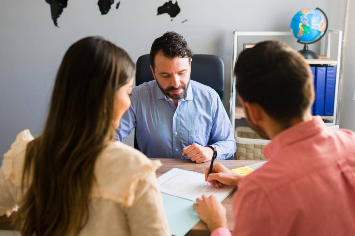 A group meeting in an office setting, with a man in a blue shirt discussing documents with two people seated at a table.