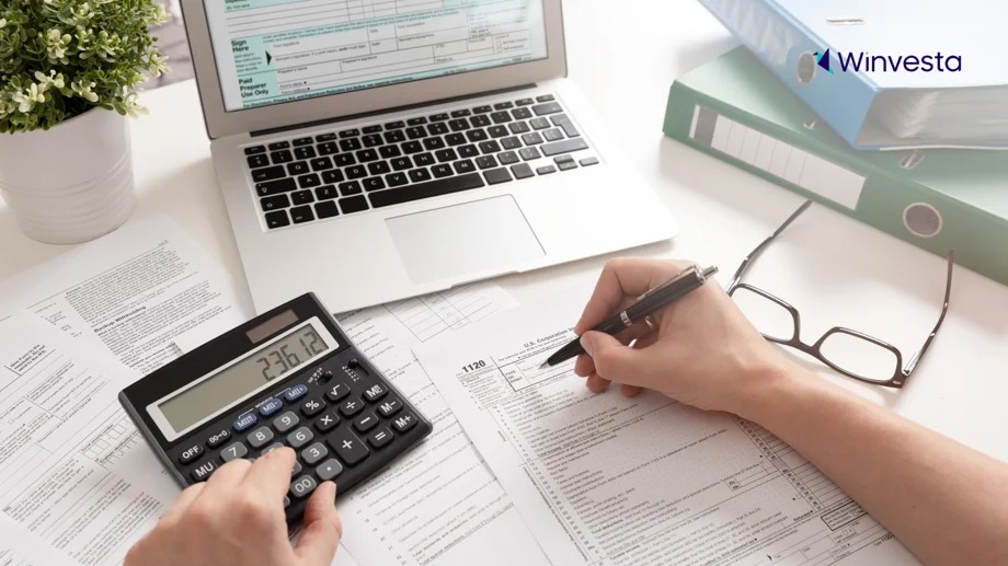 A person calculates numbers on a calculator while filling out tax forms, with a laptop and folders in the background.