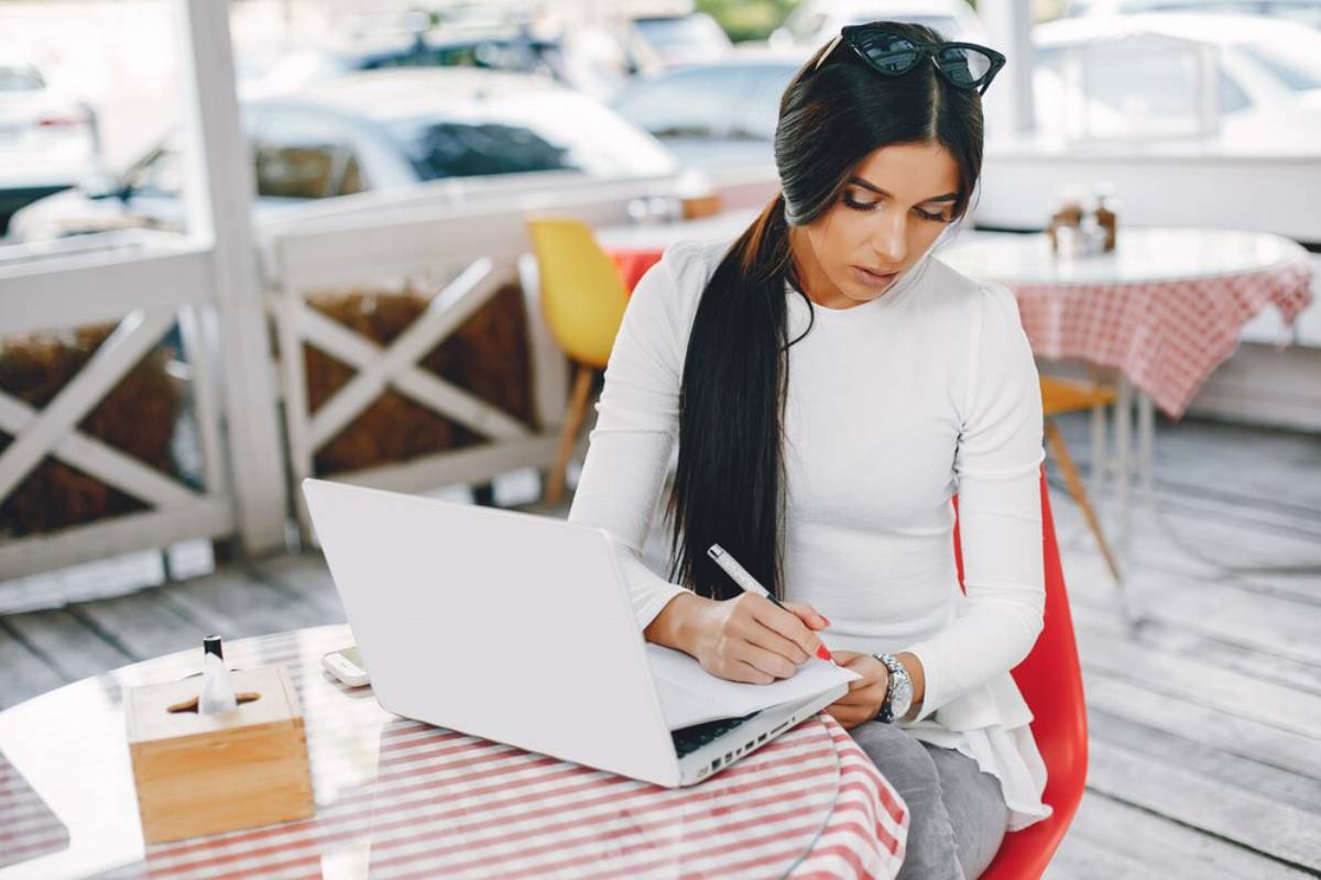 A woman sits at a café table with a laptop open, jotting notes with a pen, dressed in a stylish white top and gray pants.