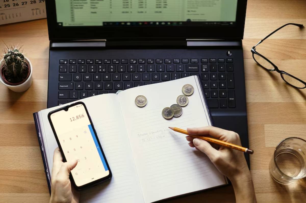 A person uses a phone calculator while jotting notes in a notebook, next to coins, a laptop, a plant, and a glass of water.