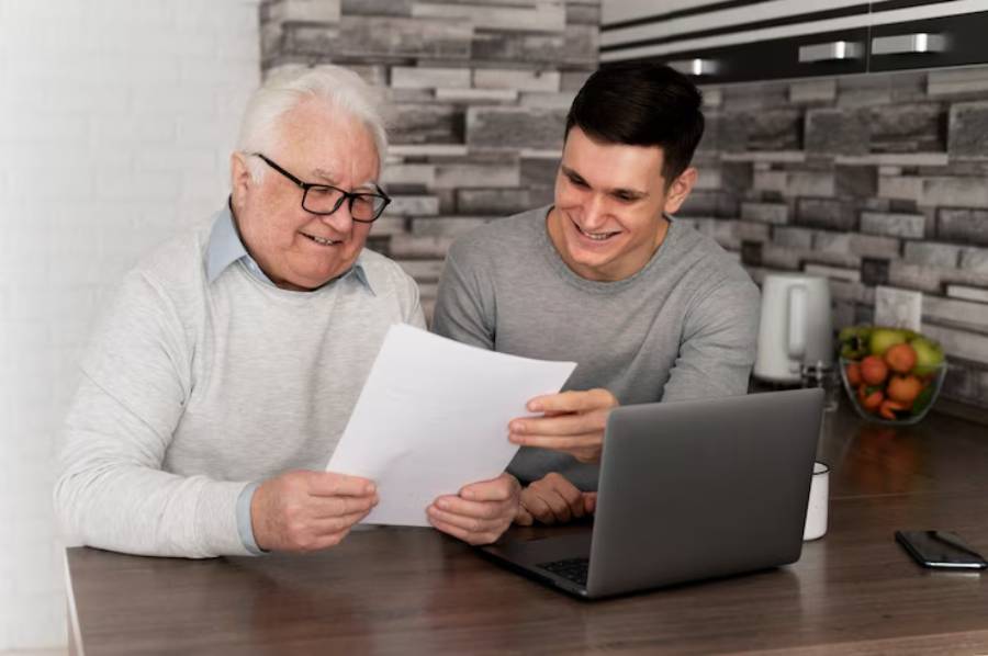An elderly man and a young man sit at a table, reviewing documents together with a laptop and fruit bowl in the background.