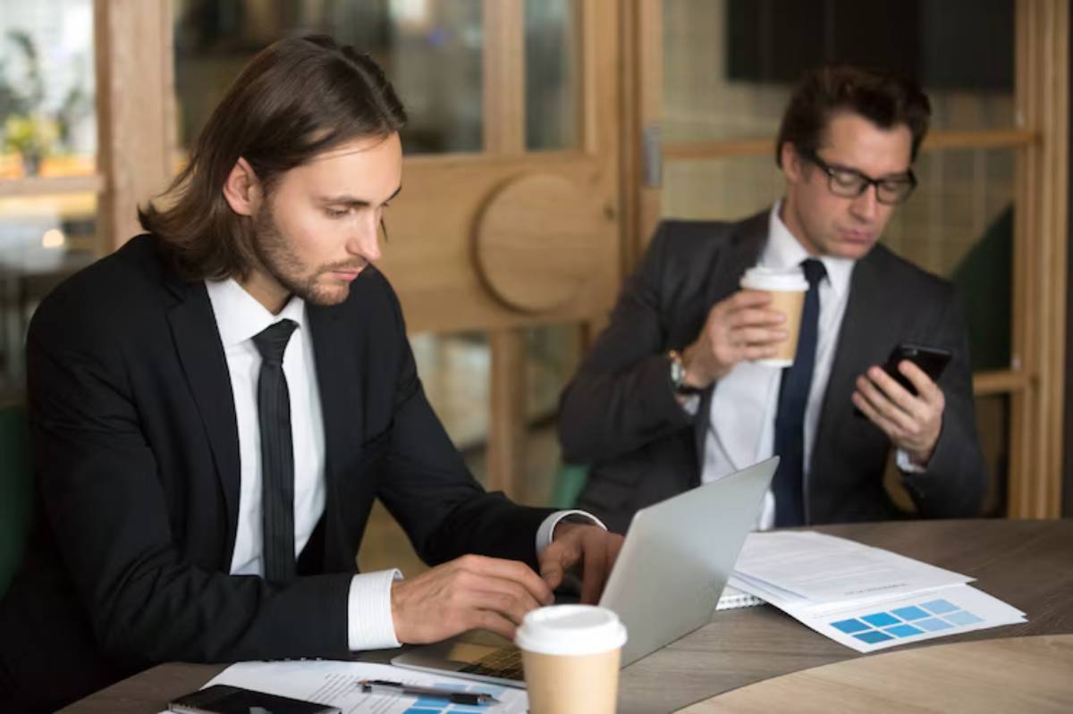Two business men in suits engage in conversation at a wooden table, one using a laptop, the other holding a coffee and a phone.