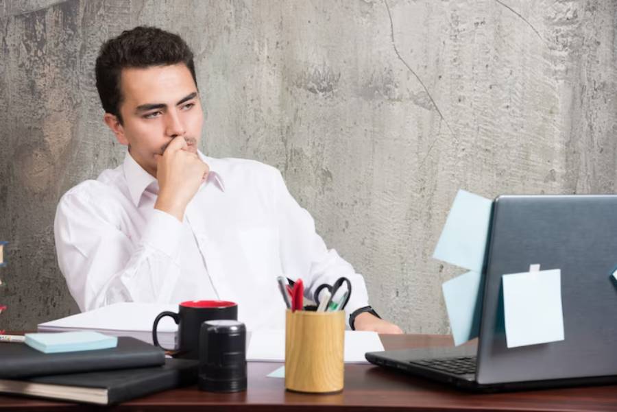 A man in a white shirt sits thoughtfully at a desk cluttered with books, stationery, and a laptop against a concrete wall.