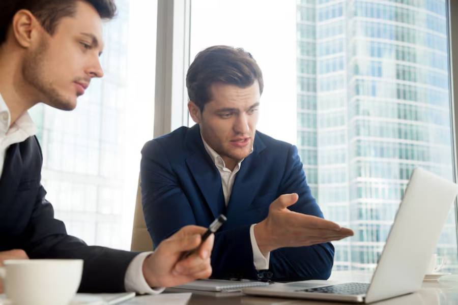Two professionals in a meeting, one gesturing with a pen while discussing a project, with a laptop and city skyline visible in the background.