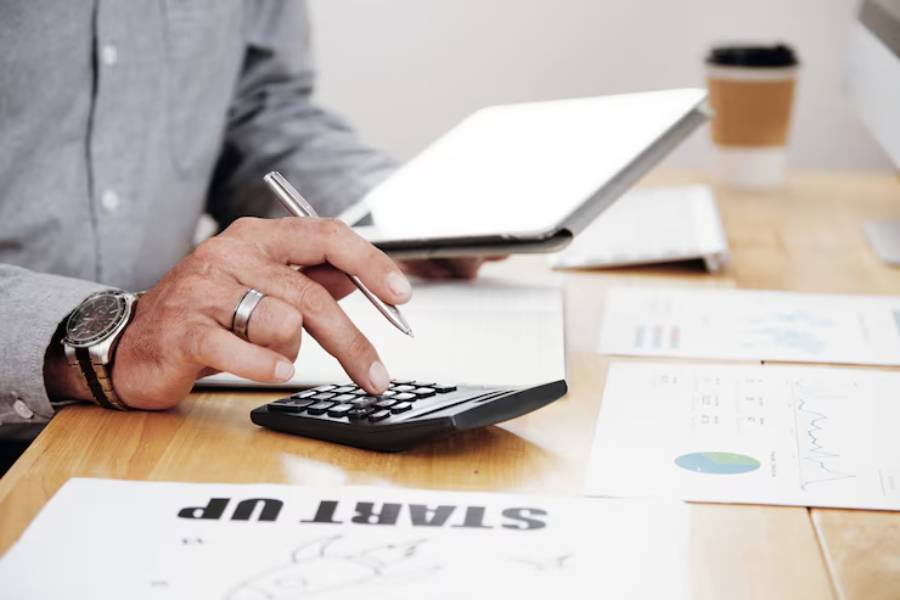 A person calculates expenses using a calculator and tablet, surrounded by documents and a coffee cup on a wooden desk.